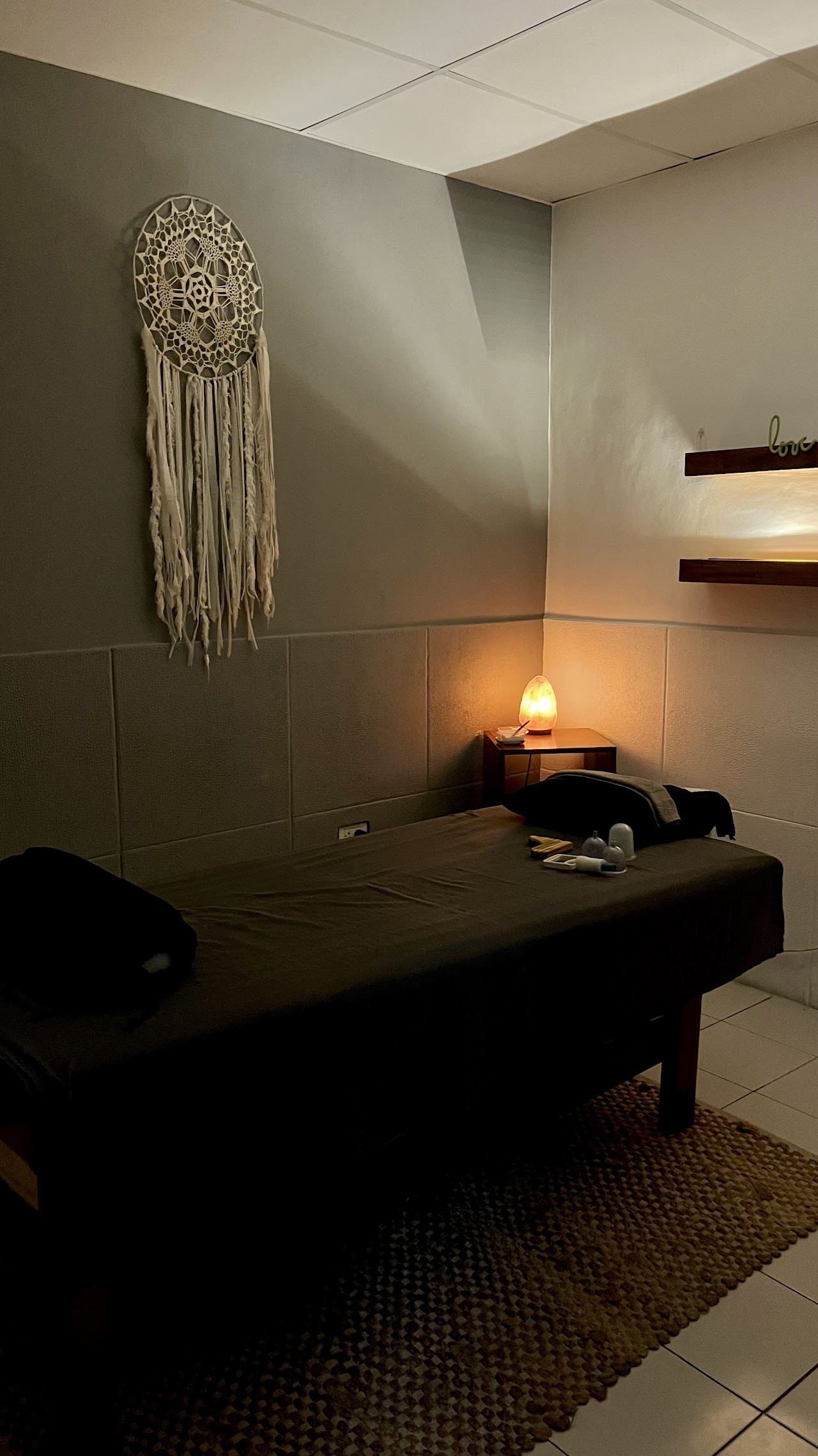 Warm cozy massage therapy room, dimly lit with warm candlelight, glowing Himalayan salt lamp, macrame dreamcatcher and woven bohemian wall decor, massage table with soft linen and neatly folded natural towels, tranquil boho-zen wellness spa retreat atmosphere, professional interior photography, soft warm ambient lighting, highly detailed.